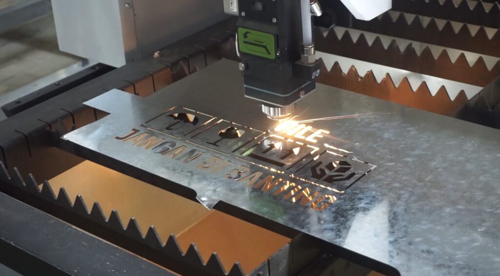 Mesin laser cutting plat besi berwarna putih dengan meja kerja luas sedang memotong lembaran plat logam. Terlihat sinar laser merah yang memotong dengan presisi di ruang workshop yang profesional.