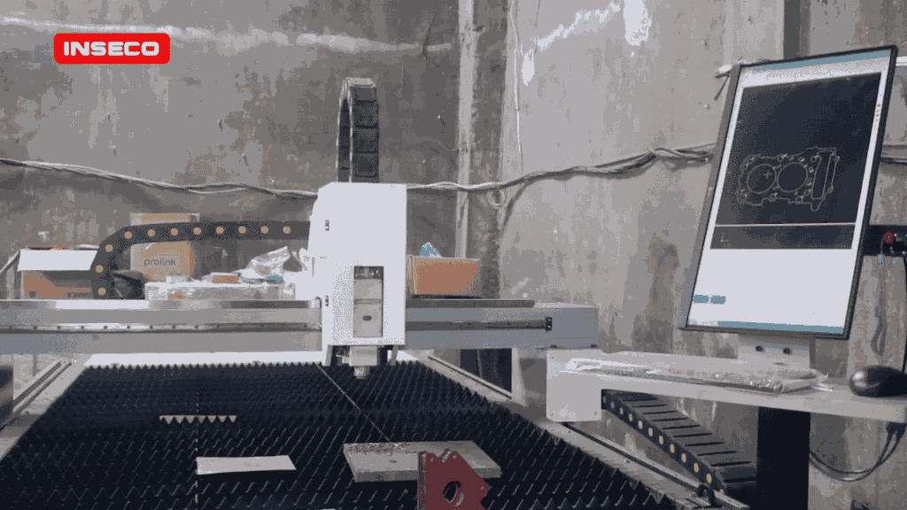 Mesin CNC fiber laser INSECO di dalam bengkel; kepala laser berada di atas meja sarang lebah, monitor samping menampilkan gambar teknik potongan logam